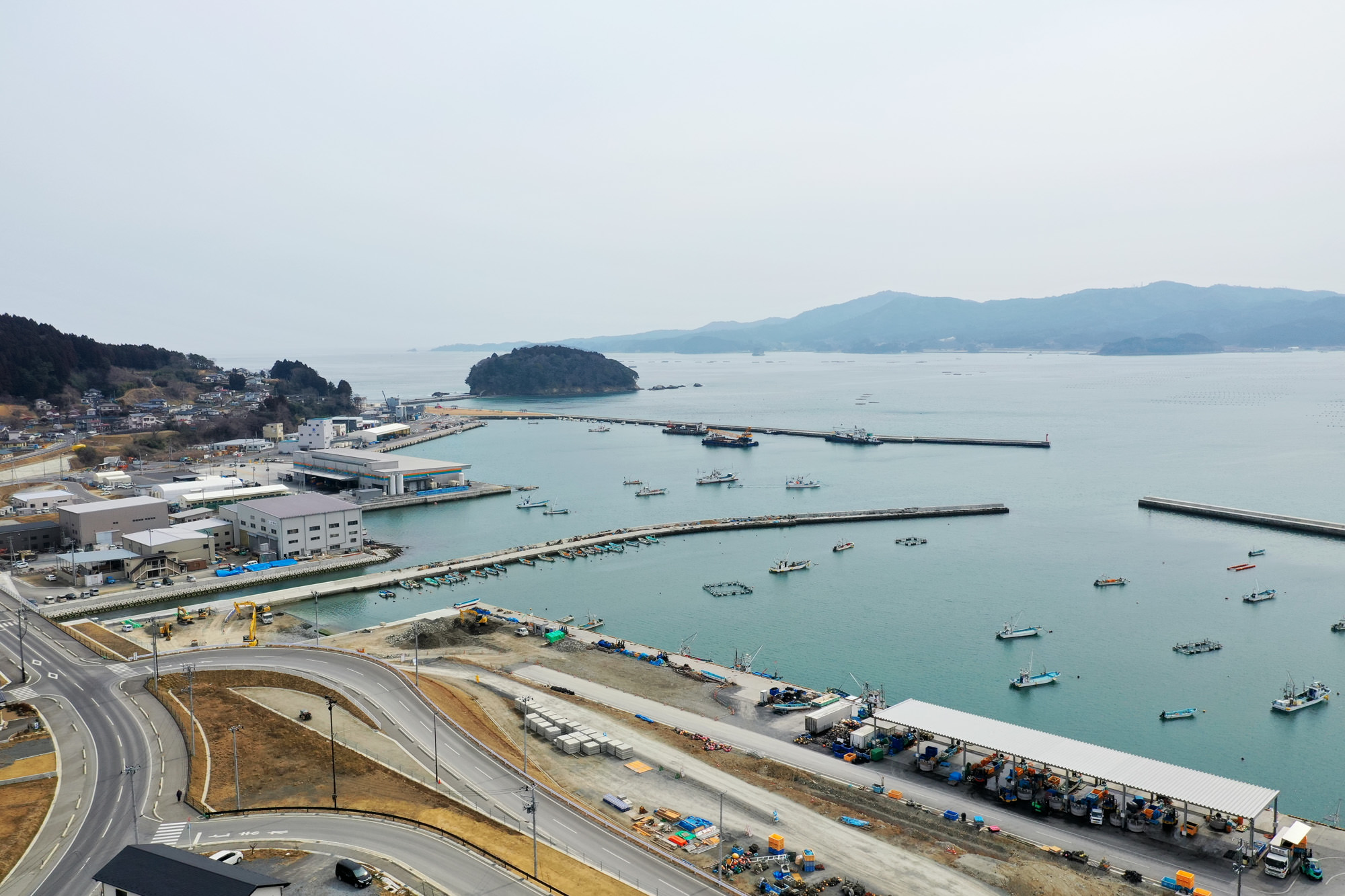 穏やかな海と曇り空の下、複数の船が停泊する港とその周辺に広がる建物や道路、工事現場、小島、そして遠くに連なる山々を上空から捉えた沿岸地域の写真