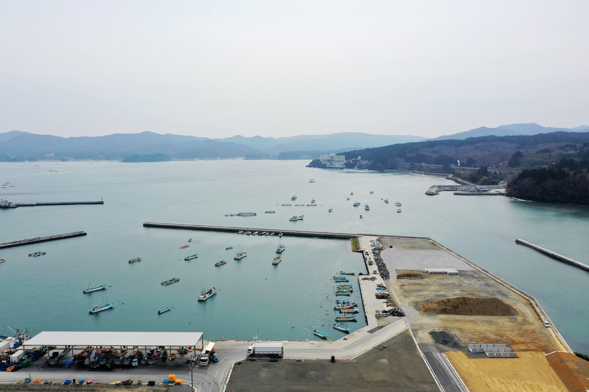 防波堤に囲まれた港に多数の小型船が停泊し、複数の桟橋が海に伸びる中、左側には建物と車が見え、背景には山や丘が広がる海辺の町を上空から捉えた港町の写真