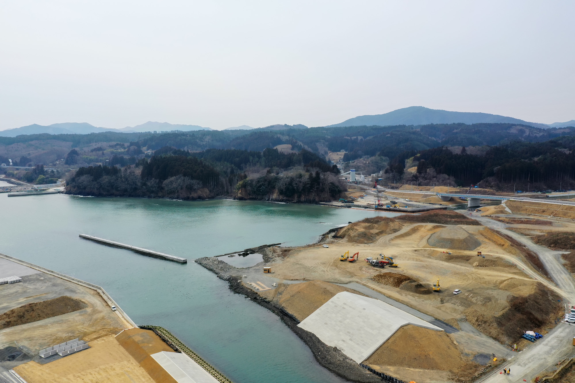 海に面した土地で重機や資材が並ぶ工事現場が広がり、周囲には木々や草地が点在し、背景には山々が連なる中、護岸工事や埋立整備が進められている様子を上空から捉えた沿岸整備の写真