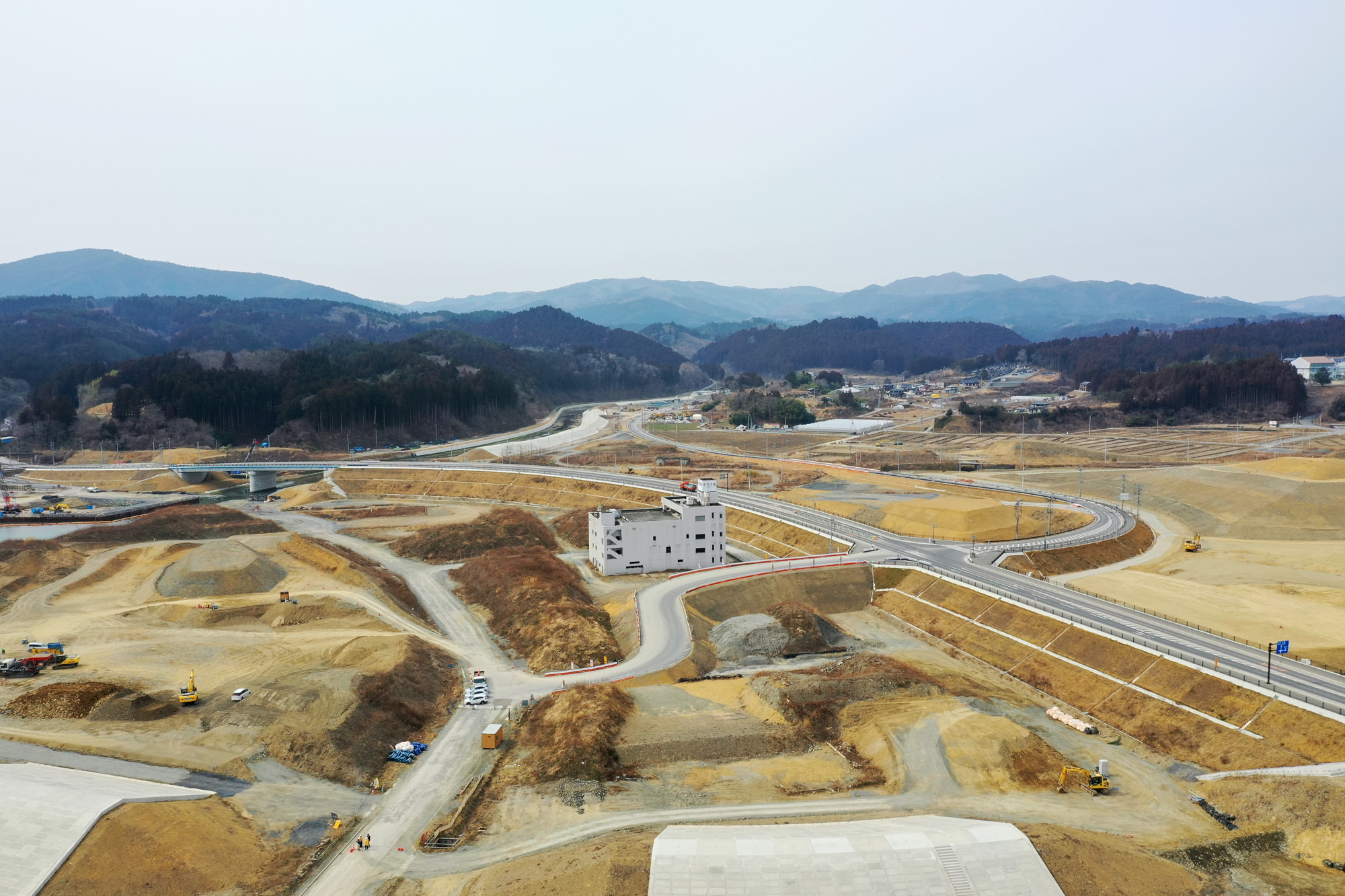 中央に建物があり、その周囲で複数の道路や通路が整備され、土の盛り上がりや建設車両が点在する広大な工事現場を上空から捉え、背景にはなだらかな丘と山々が広がる造成地の写真