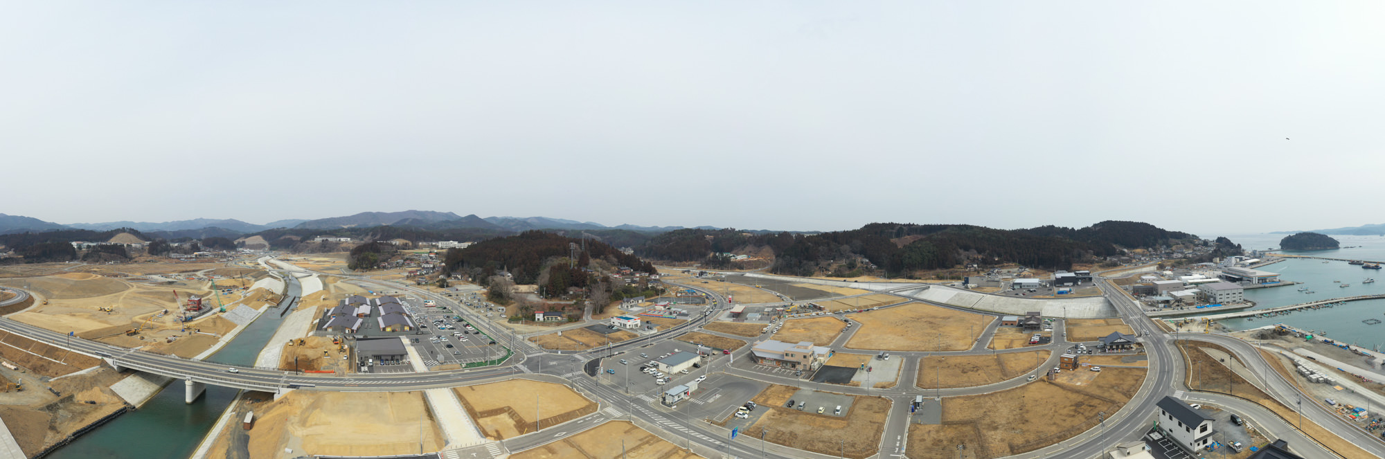 海や川に囲まれた沿岸の町を広く見渡す構図で、複数の道路や橋、建物が点在し、周囲の丘陵に囲まれながら一部地域では新たな建設や再整備が進められている様子を捉えた沿岸都市の写真