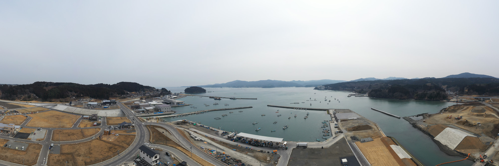 海に面した港湾地域を上空から捉えた構図で、複数の桟橋や埠頭に多くの船が停泊しており、周囲には建物や道路、工事現場が広がり、背景には山々が連なる開発と自然が混在する港町の写真