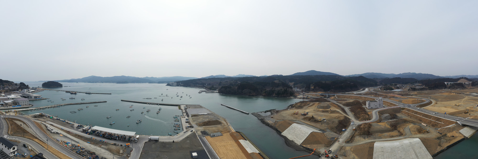 港に複数の船や桟橋が並び、周囲の陸地では道路や建物の整備が進み、広い造成地が広がる中、遠くには山々が連なり、晴れた空の下で開発が進む沿岸地域を上空から捉えた港湾開発の写真