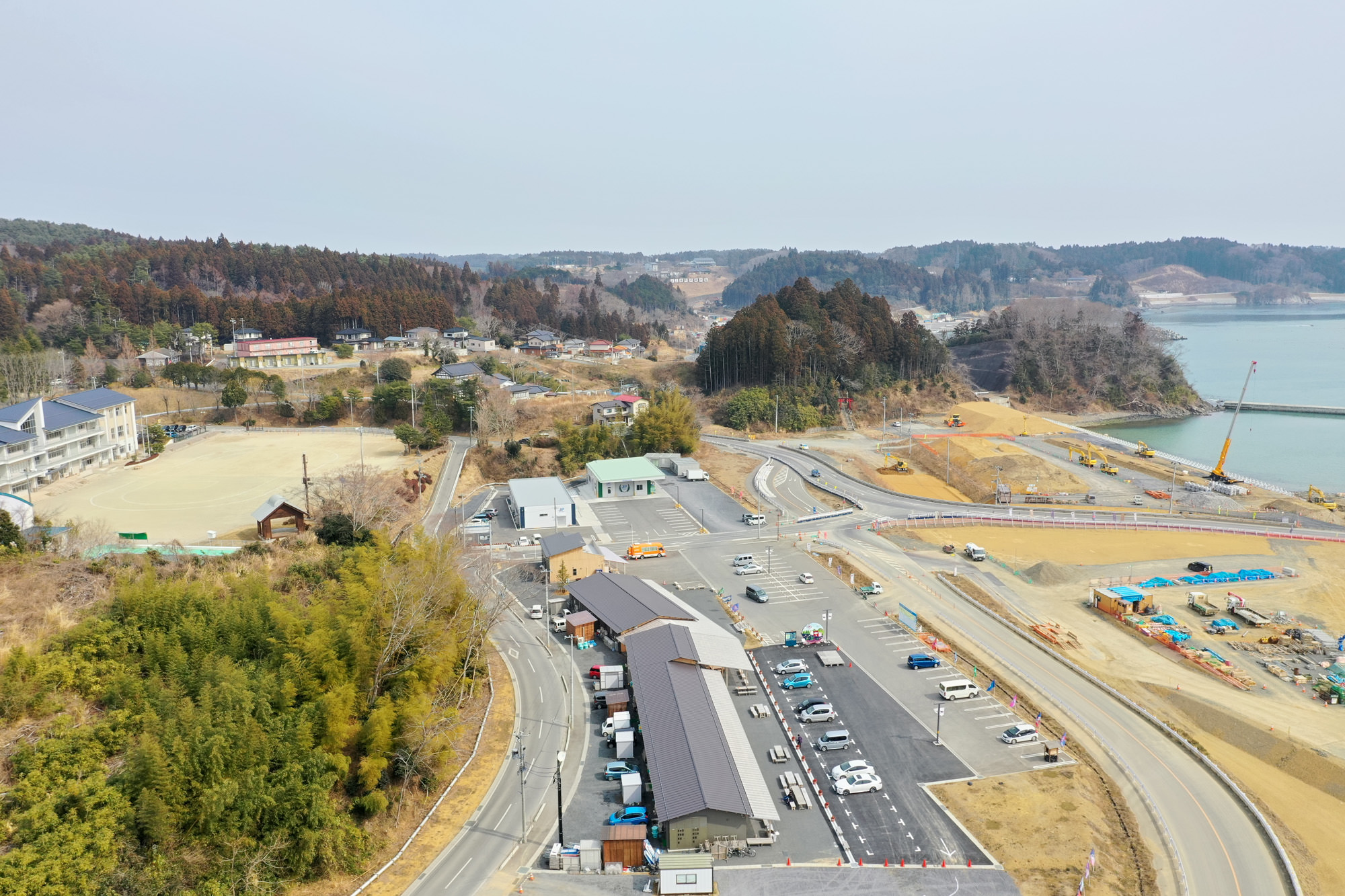 海岸線を背景に、建物や駐車場、道路が点在し、森林や草地、海沿いの工事現場が広がる自然と開発が混在する地域を上空から捉えた沿岸地域の写真