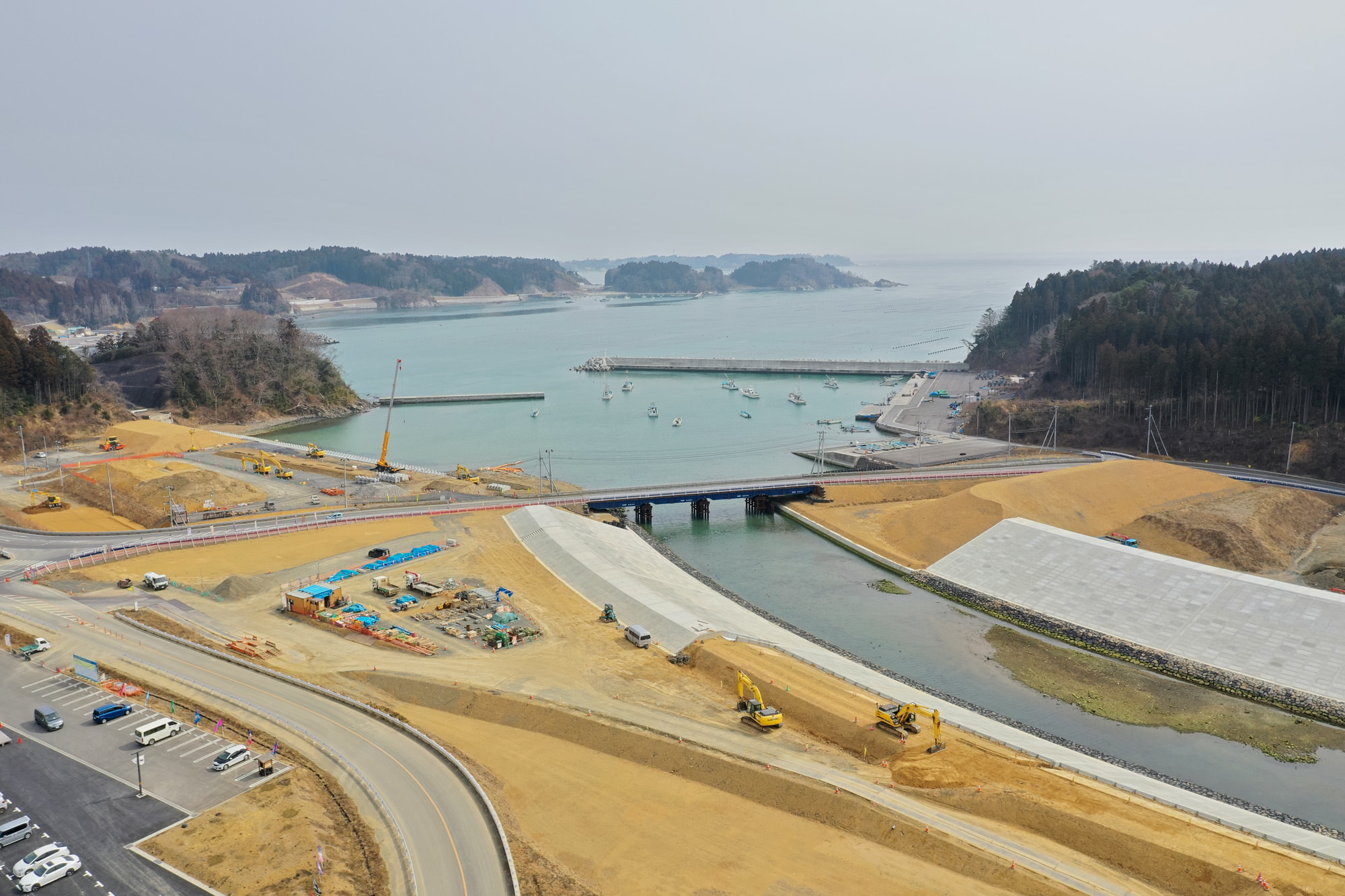海へと続く水路にかかる橋を中心に、重機やクレーンが稼働する沿岸の工事現場が広がり、周囲には丘や木々があり、背景には船が停泊する海が見える、海岸近くでのインフラ整備の様子を上空から捉えた沿岸工事の写真