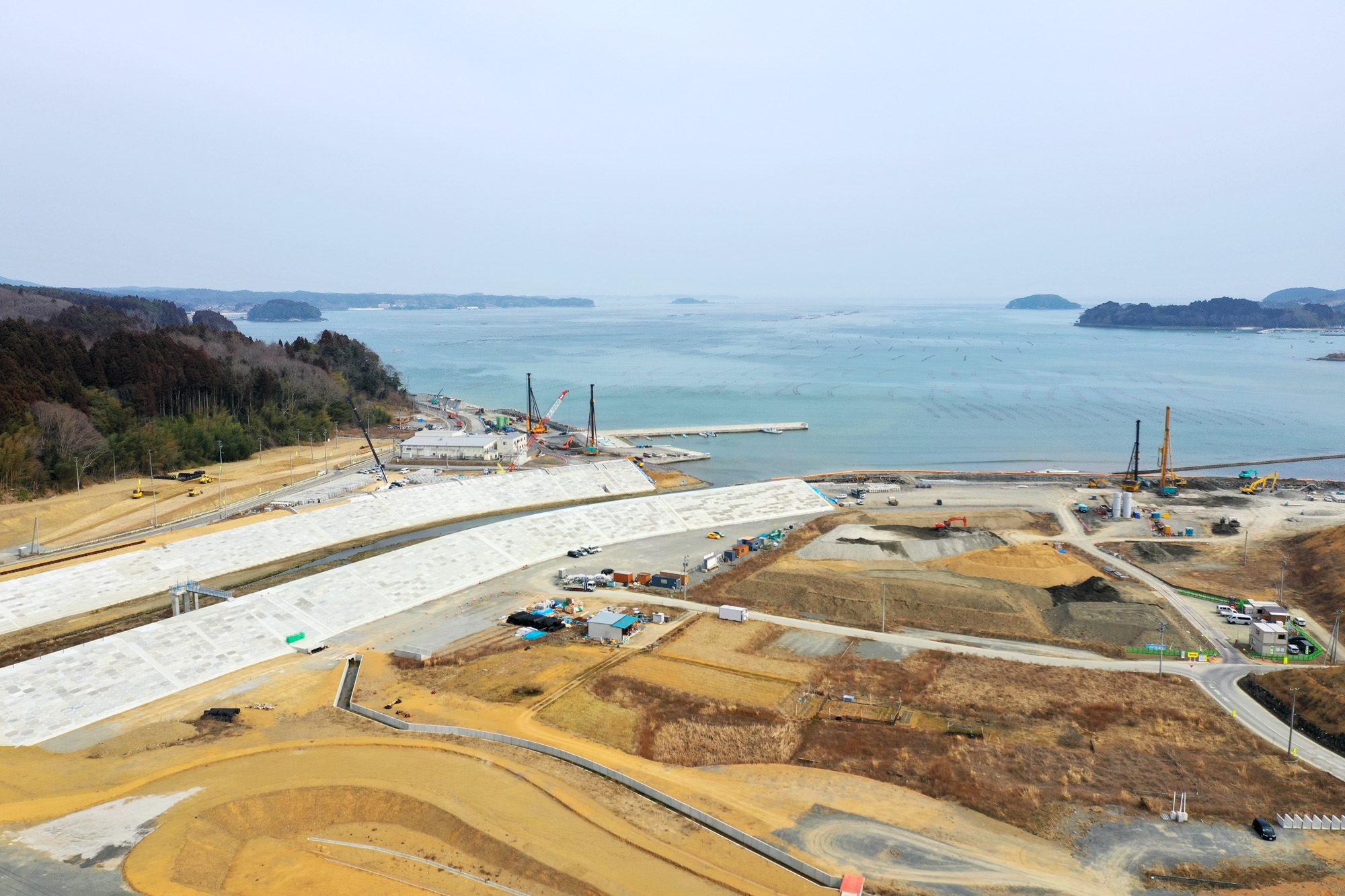 海岸沿いの工事現場で、白い大型構造物が海岸線に沿って建設されており、周囲には複数のクレーンや重機、仮設施設や車両が点在し、背後には海と小さな島々が広がる海辺の開発風景の写真