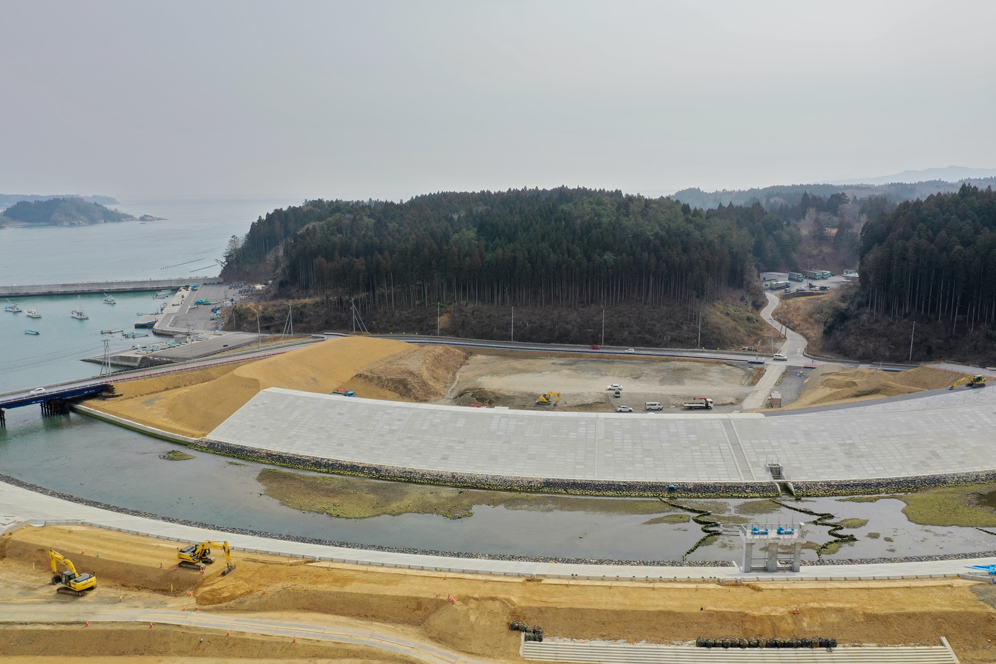 森林に囲まれた海辺の工事現場で、複数の重機が稼働し、コンクリート構造物や土砂が広がる中、道路が現場へと続いている様子を上空から捉えた沿岸整備の写真