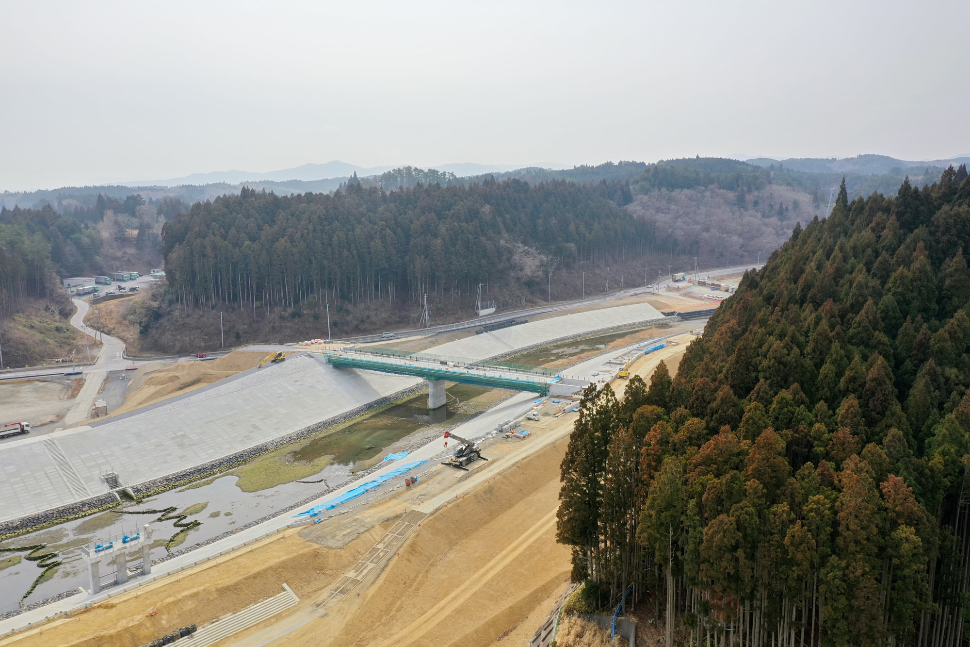 森林に囲まれた農村地帯に新しく架けられた橋が川や運河を横断し、周囲には建設資材や機械が点在するインフラ整備中の様子を上空から捉えた山間部の橋梁工事の写真