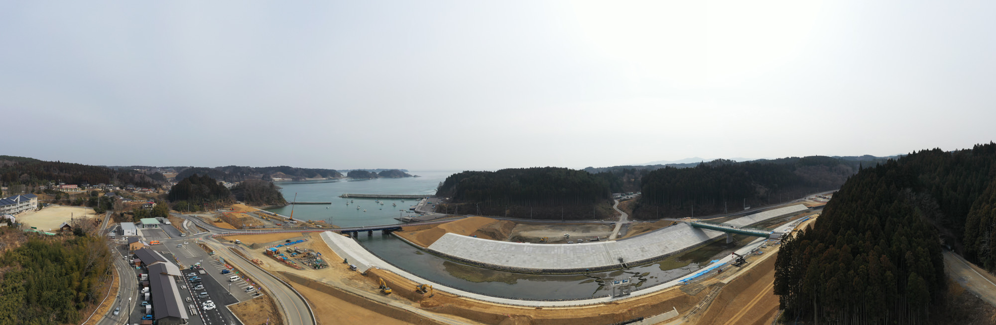 入り江のような海を囲む森林と丘陵の中に広がる大規模な工事現場で、橋が水域を横断し、道路や駐車場、建設中の構造物が点在し、重機や車両が稼働する様子を曇り空の下で上空から捉えた沿岸開発の写真