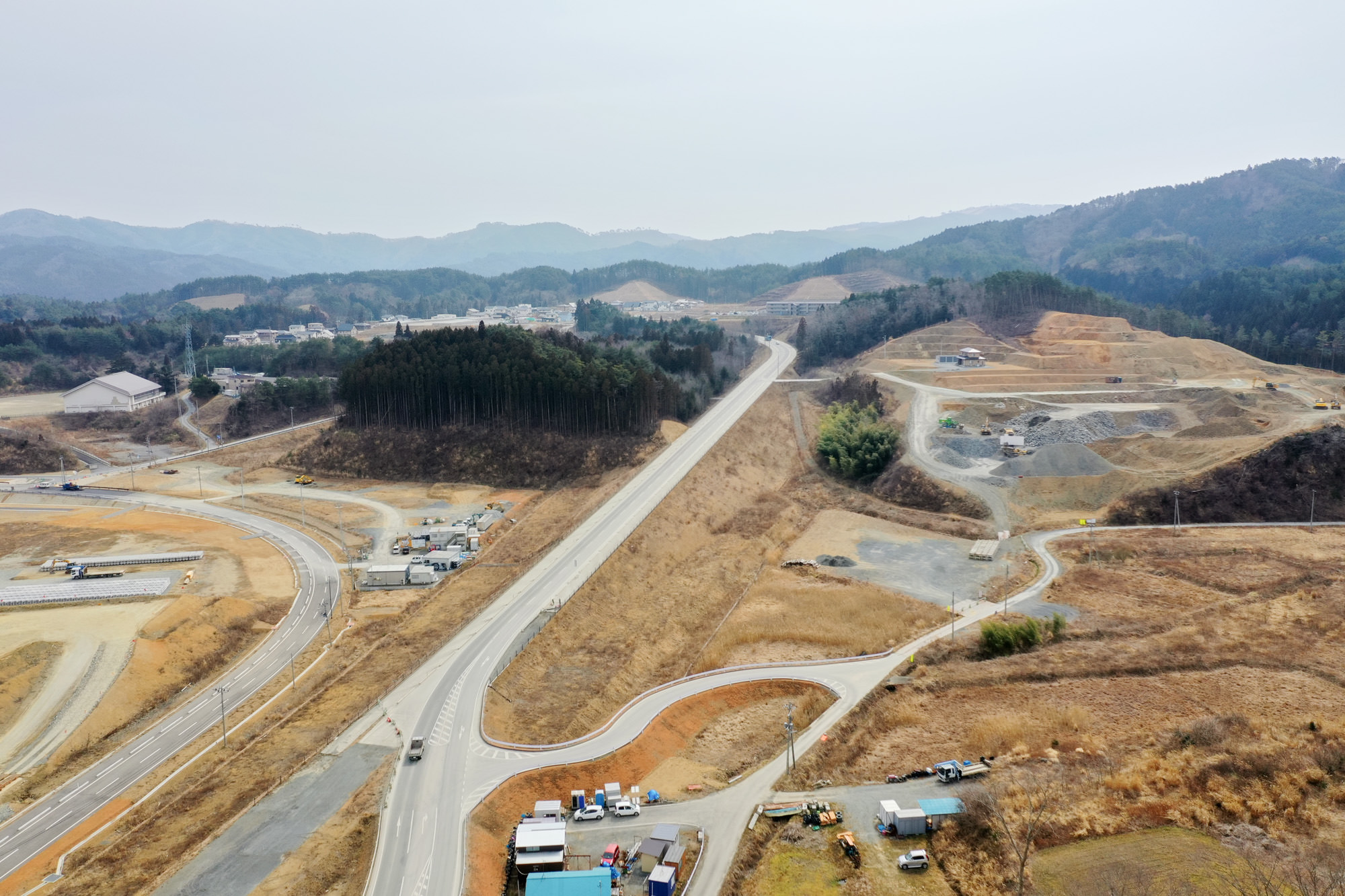 空から見下ろした農村地域の風景で、曲がりくねった道路や点在する建物、森林の一部が見られ、背景には丘が広がり、所々に工事や掘削の跡が確認できる乾いた土地の写真