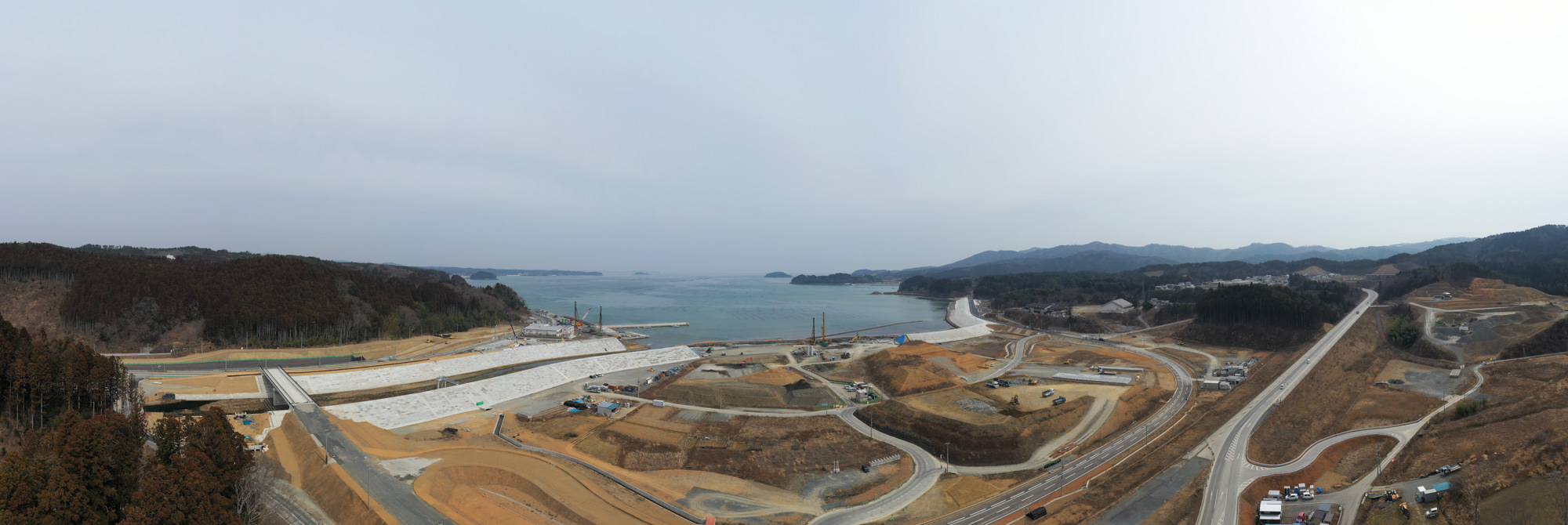 海に面した工事現場を広く見渡す風景で、周囲には丘や森林が広がり、背景には海が見え、敷地内には曲がりくねった道路や通路、重機や資材が点在し、地面がむき出しの部分や建設途中の構造物が見られる開発初期段階の写真