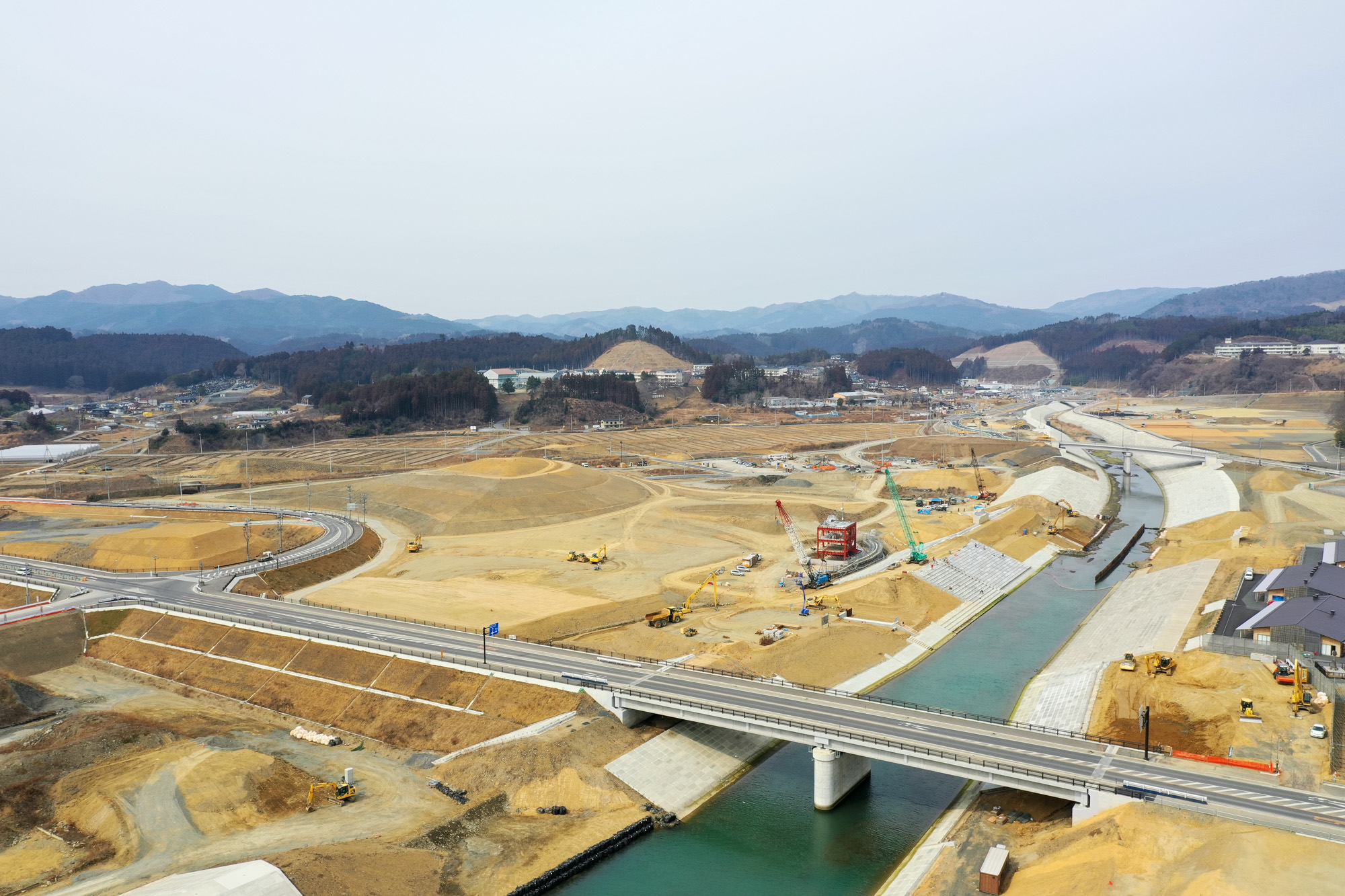 運河または川にかかる橋を中心に、周囲の山々を背景に広がる大規模な工事現場で、クレーンやショベルカーなどの建設機械が稼働し、道路や堤防などのインフラ整備が進められている様子を捉えた工事現場の写真