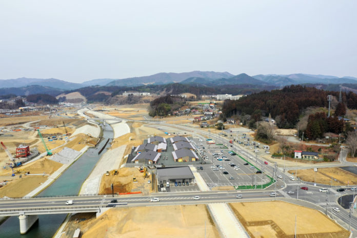 広い範囲にわたる工事現場の風景で、中央には川が流れ、その両岸には建物や駐車場、道路が整備されている様子が一望できる地域開発の写真
