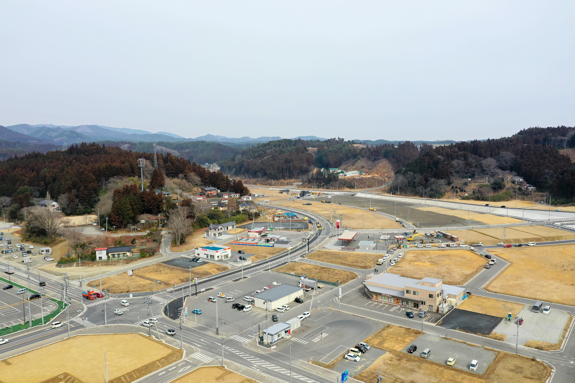 木々に覆われた丘を背景に、建物や道路、駐車場が点在する農村地域を上空から捉えた構図で、周囲には広がる畑や車が走る道路が見え、曇り空の下で静かな雰囲気が漂う地方の町の写真