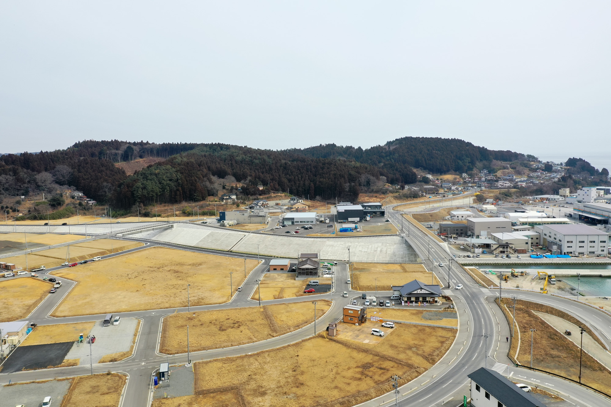 曲がりくねった道路や小さな建物、背景にある大きな施設が点在し、茶色い草地や土の空き地が広がる半都市型の地域を上空から捉え、遠くには森林に覆われた丘が見え、曇り空の下で静けさを感じさせる地域開発の写真