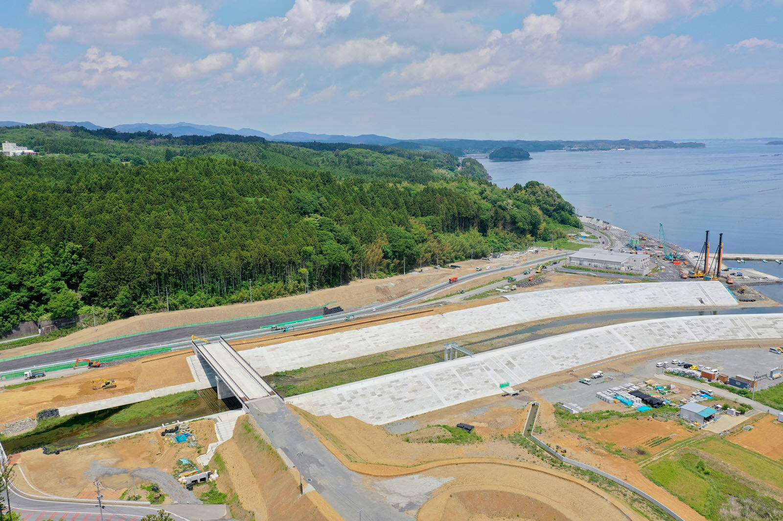 海沿いに新しく建設されたインフラ施設があり、大きなコンクリートの構造物が海岸線に沿って延び、周囲には工事用の機材や資材が点在し、背景には密集した森林の丘と海が広がり、空は晴れている防潮堤の写真