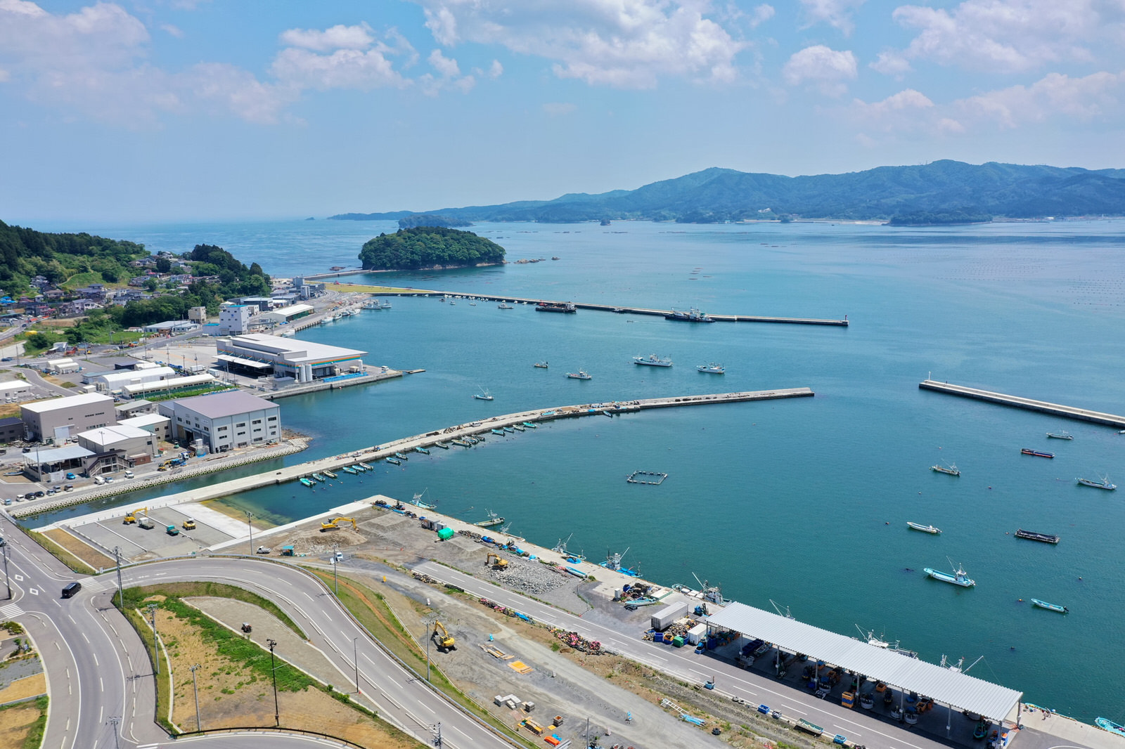 港のある海沿いの地域を上空から撮影した写真で、水面には複数の桟橋や船があり、海岸線には建物や道路が並び、遠くには小さな島が見え、周囲には丘や山々が広がり、空には雲が浮かぶ海辺の港の写真