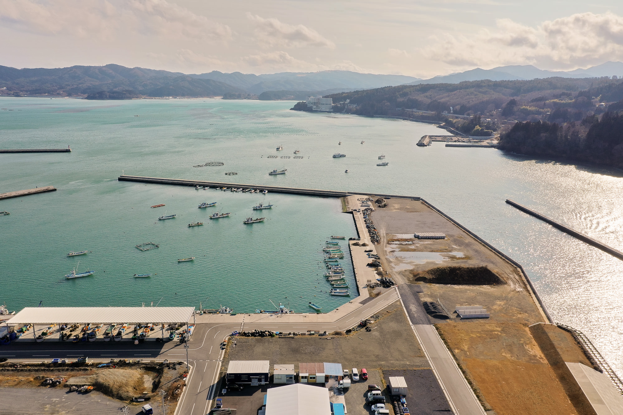 海沿いの地域を空から撮影した写真で、港には複数の船が桟橋や防波堤に沿って停泊しており、海岸近くには建物や構造物、道路や駐車場が広がり、背景には丘や山々が見え、空には雲が少し浮かぶ写真