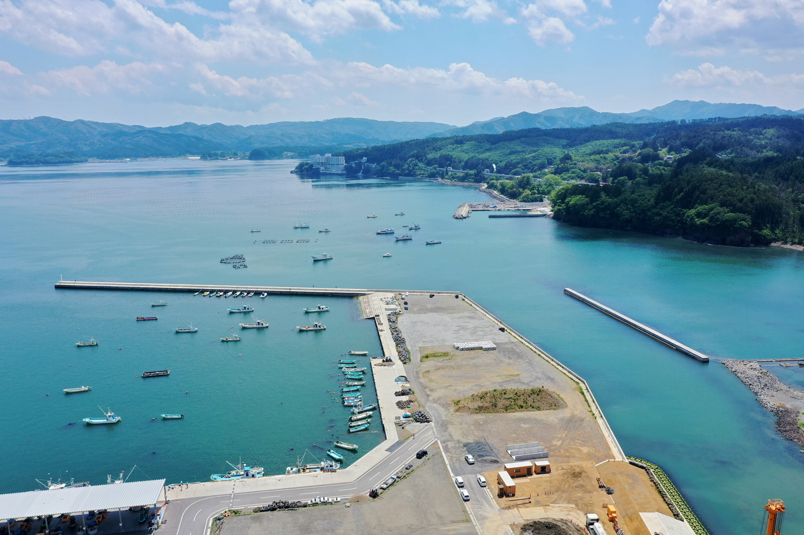 穏やかな水面に複数の船が停泊する港を中心に、海岸線には建物や工事現場が点在し、周囲には緑豊かな丘や山々が広がり、空には雲と青空が見える海辺の港の写真