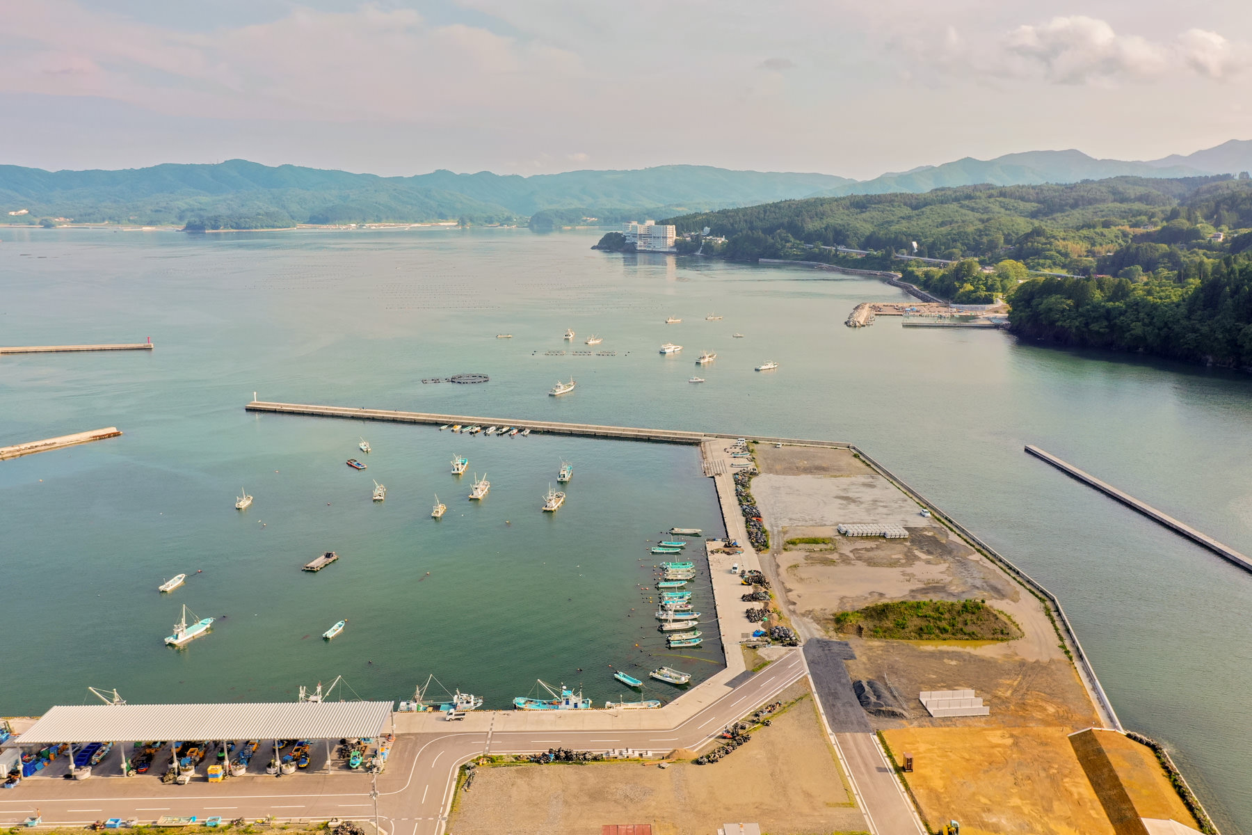 防波堤に囲まれた港に複数の船が停泊している海辺の地域を上空から見下ろした風景で、緑地と開発された区域が混在し、遠くには山々が広がっている写真