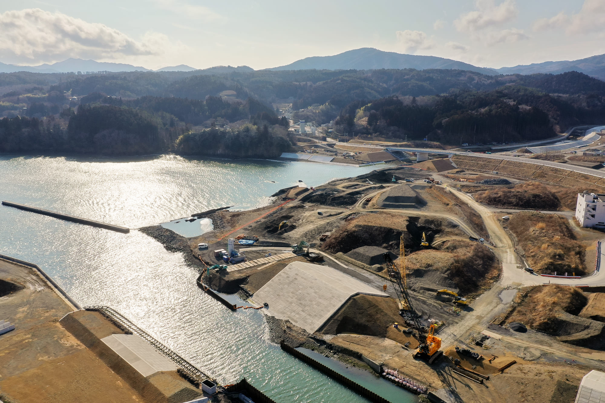 海沿いの工事現場を空から撮影した写真で、重機や構造物が点在し、水面が太陽光を反射して輝き、周囲には丘陵地や道路が広がり、背景には山々と点在する建物が見える写真