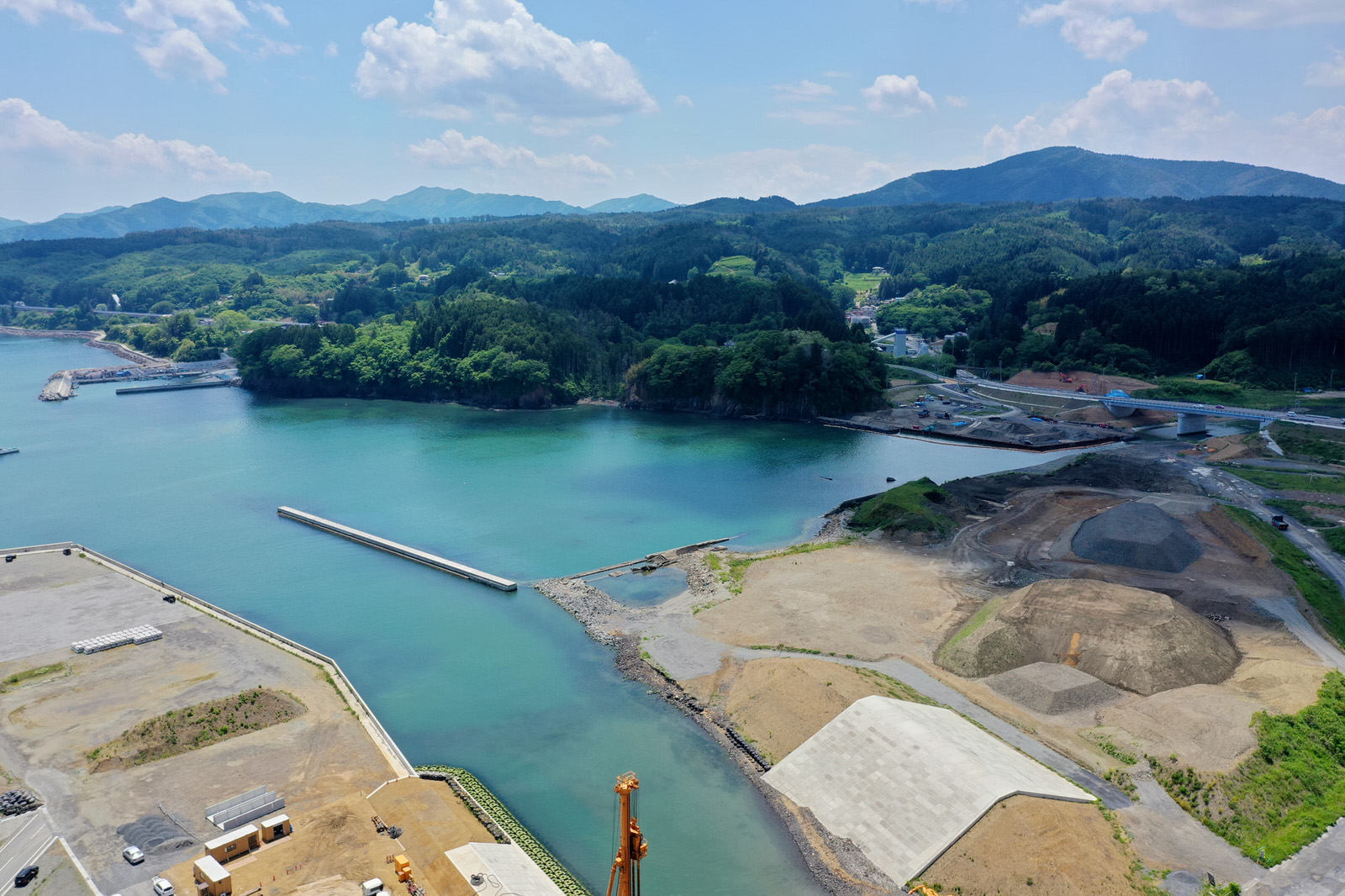 海に面した地域を上空から撮影した写真で、水辺を囲む陸地には工事現場が広がり、土がむき出しの場所やコンクリート構造物が点在し、背景には緑豊かな丘や山々が広がり、空には雲が浮かぶ海岸開発地域の写真