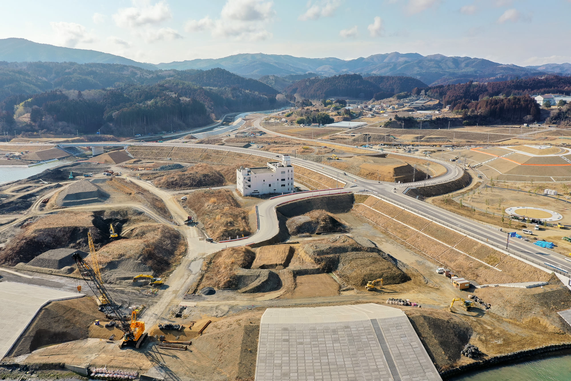 山に囲まれた丘陵地帯の工事現場を空から撮影した写真で、複数の道路や小道が走り、白い建物が中央に位置し、周囲には土の盛り上がりや造成中の区画が広がり、建設車両や機材が点在する写真