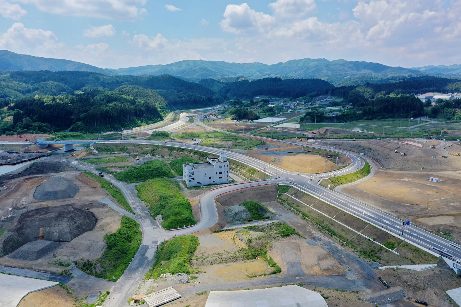 山々を背景にした農村地域で、複数の道路や小道が整備され、いくつかの構造物と緑地が点在する大規模な建設現場を上空から撮影したインフラ整備中の地域の写真