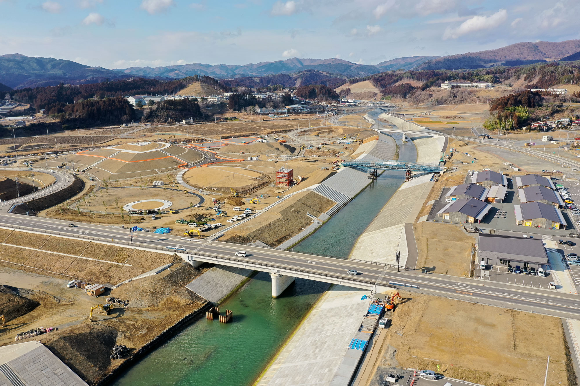 川が流れる広大な工事現場を空から撮影した写真で、川には橋が架かり、周囲には建設車両や機材が点在し、右側にはいくつかの建物があり、背景には丘や山々が広がる写真