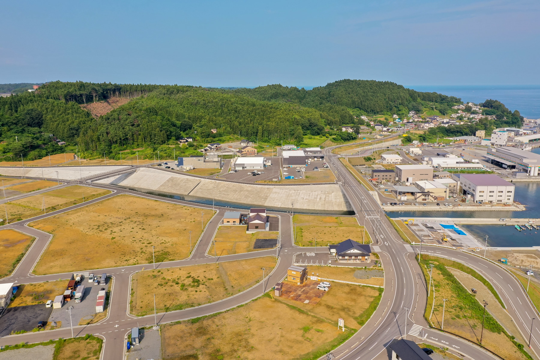 海に面した町を上空から見下ろした風景で、道路や建物が整然と配置され、未開発の空き地も点在しており、背景には森林に覆われた丘が広がっている写真
