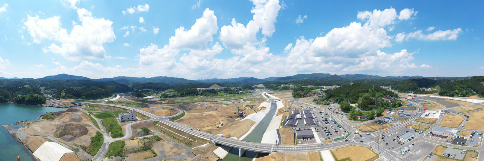 中央に川が流れ、その上に複数の橋が架かり、周囲には道路や建物、駐車場、工事現場が点在し、背景には丘や山々が広がり、空には雲が浮かぶ自然と都市開発が混在する地域の写真