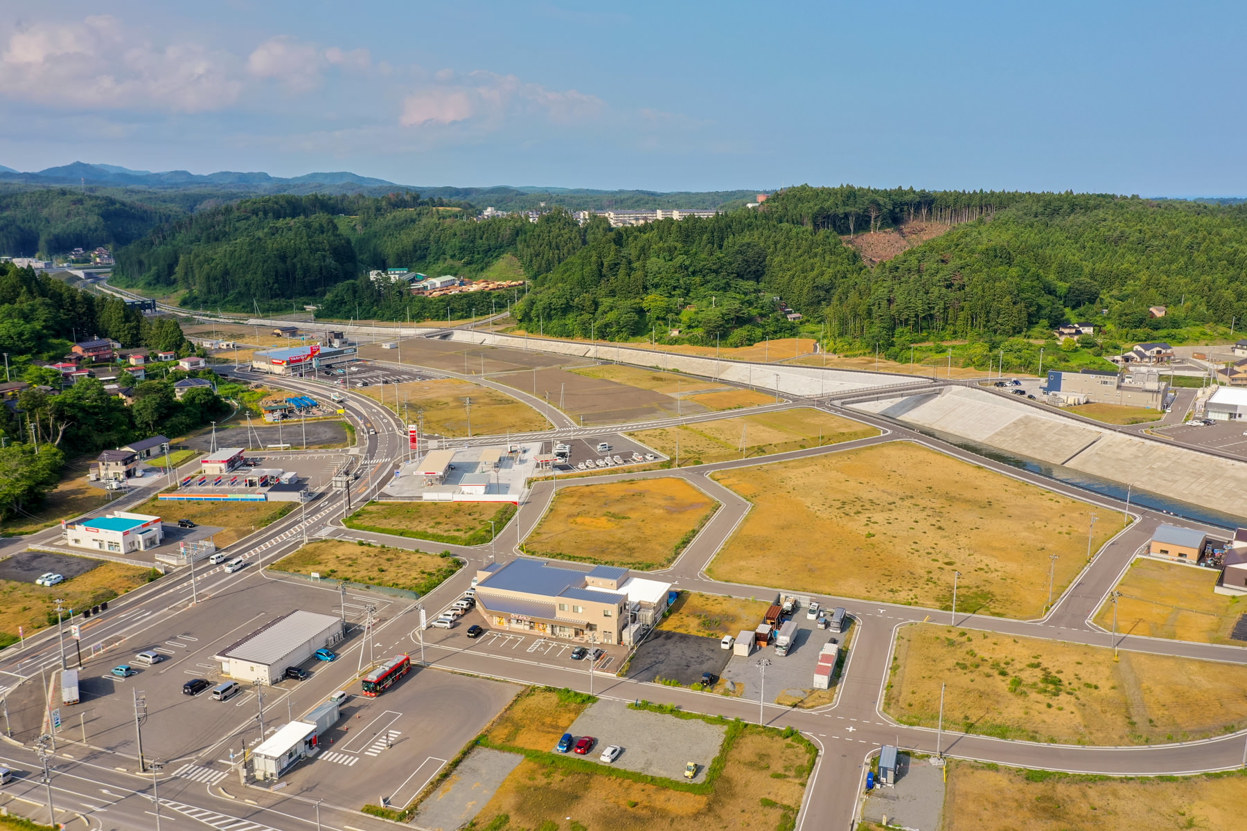 緑に覆われた山々に囲まれた地域に、広々とした空き地と整備された道路が広がっており、周囲には建物が点在している町の写真