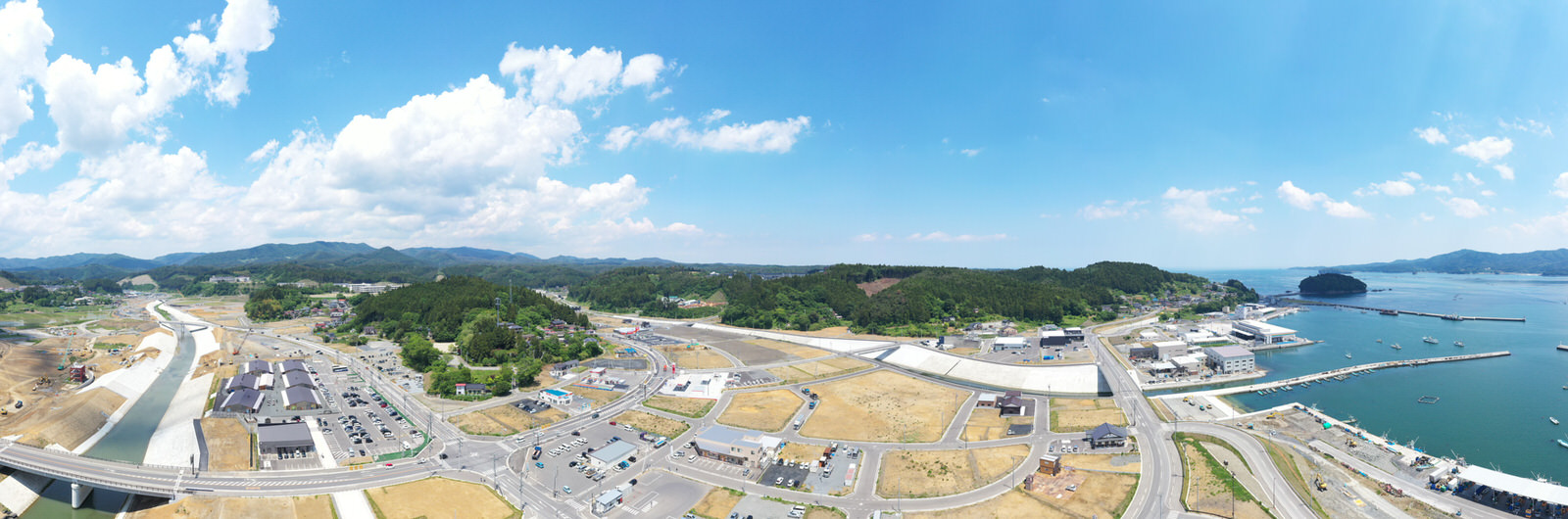 海に面した町を広く見渡す写真で、町にはさまざまな建物や道路が広がり、周囲には緑と丘陵地が広がっていて、水面には複数の船が浮かび、空には雲が点在する海辺の町の写真