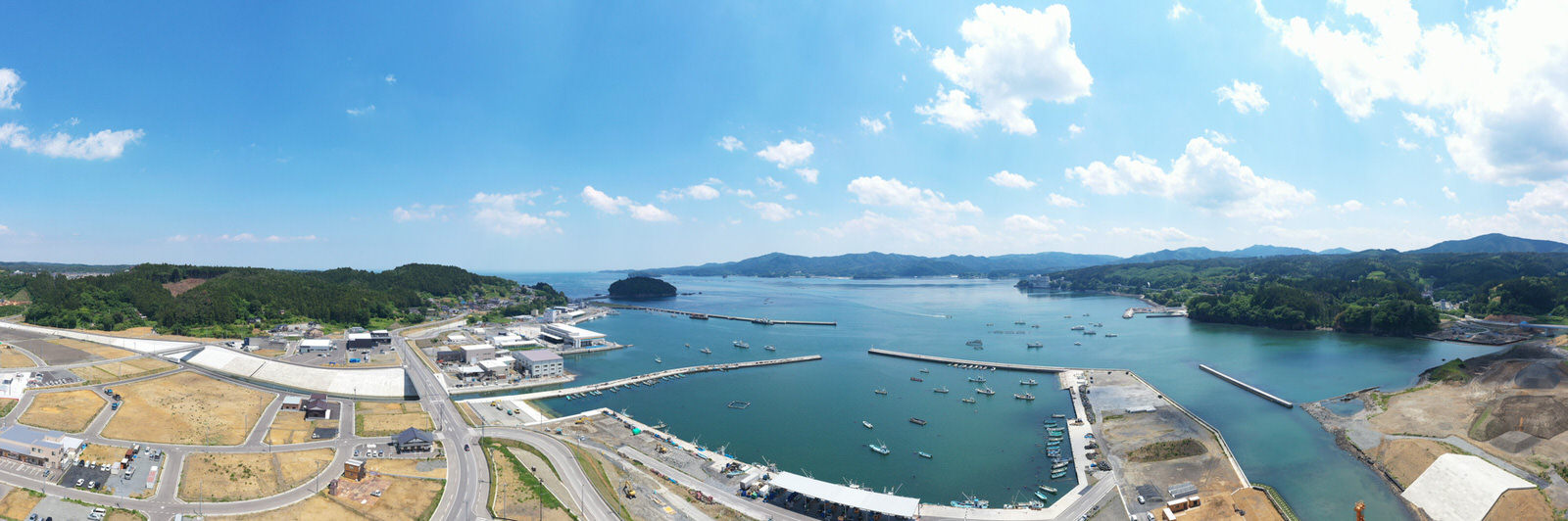 港のある海沿いの地域を広く見渡す写真で、水面には複数の船が停泊しており、海岸線にはさまざまな建物や構造物が並び、周囲には緑の丘や開けた土地が広がり、道路が地域をつないでいて、空には雲が点在する晴れた日の写真