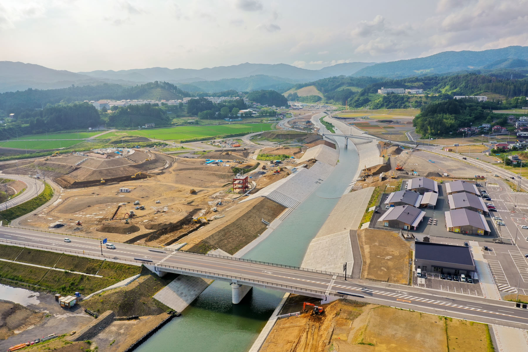 広い谷間に位置する川の上に橋が建設されており、橋の両端には重機や資材が並ぶ工事現場が広がっていて、周囲には緑豊かな山々が見える風景の写真