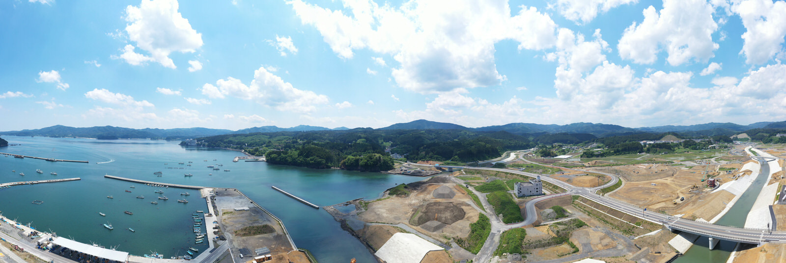 左側に複数の船が停泊する海が広がり、陸地には開発された区域と未開発の土地が混在し、右側には橋が架かっていて、背景には丘や山々が広がり、空には雲が浮かぶ海沿いの地域の写真