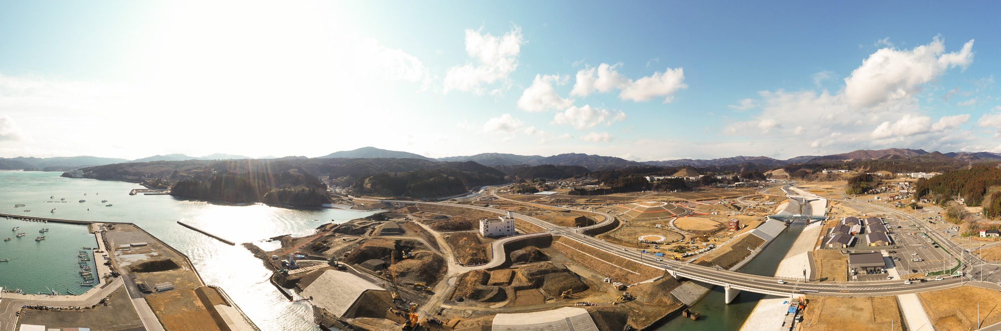 海岸沿いの広大な土地に建設中の施設や道路が広がり、背景には連なる山々が見える写真