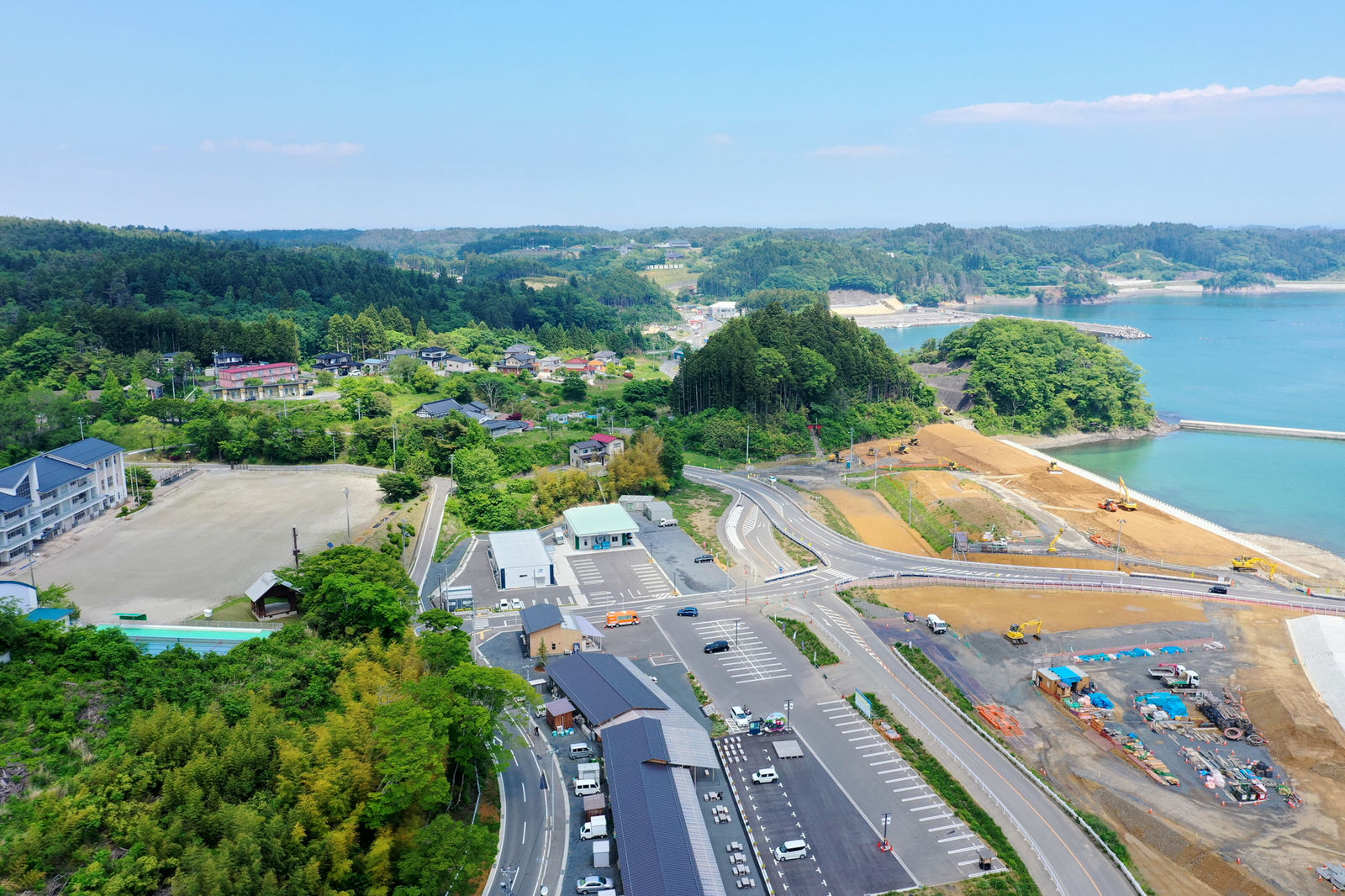 自然と開発が混在する海沿いの地域を上空から撮影した写真で、複数の建物や駐車場、道路が広がり、右側には水辺があり、その周辺では工事が行われていて、背景には緑の丘や木々が広がる海岸地域の写真
