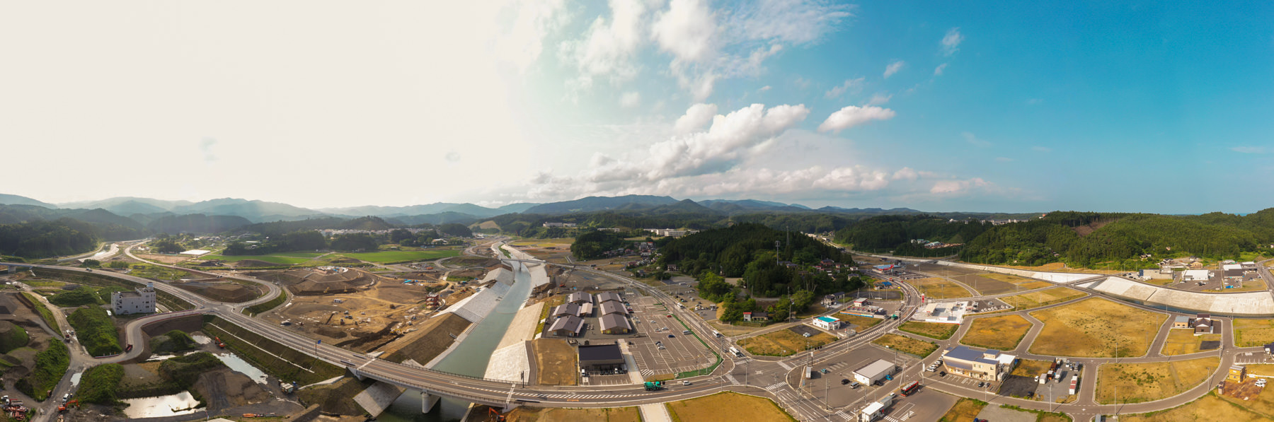 開発された土地と未開発の土地が混在する農村地域の広角風景で、中央には川が流れ、道路や建物が点在し、背景には丘や山が広がり、空には雲が部分的に浮かんでいる自然と人の営みが共存する写真
