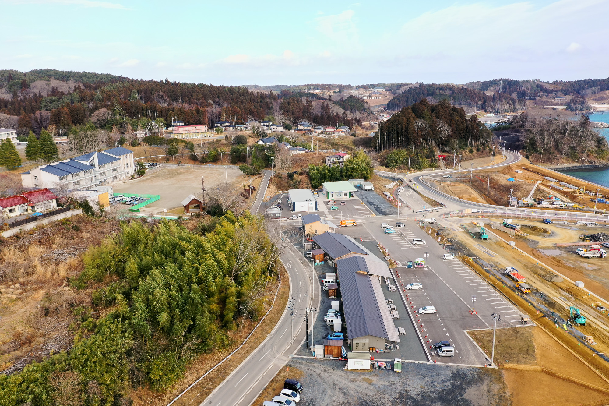 田園地帯を空から撮影した写真で、複数の住宅や大きな建物、道路、駐車場が点在し、丘陵地には木々が密集する場所と開けた土地があり、右側には水辺が広がる写真