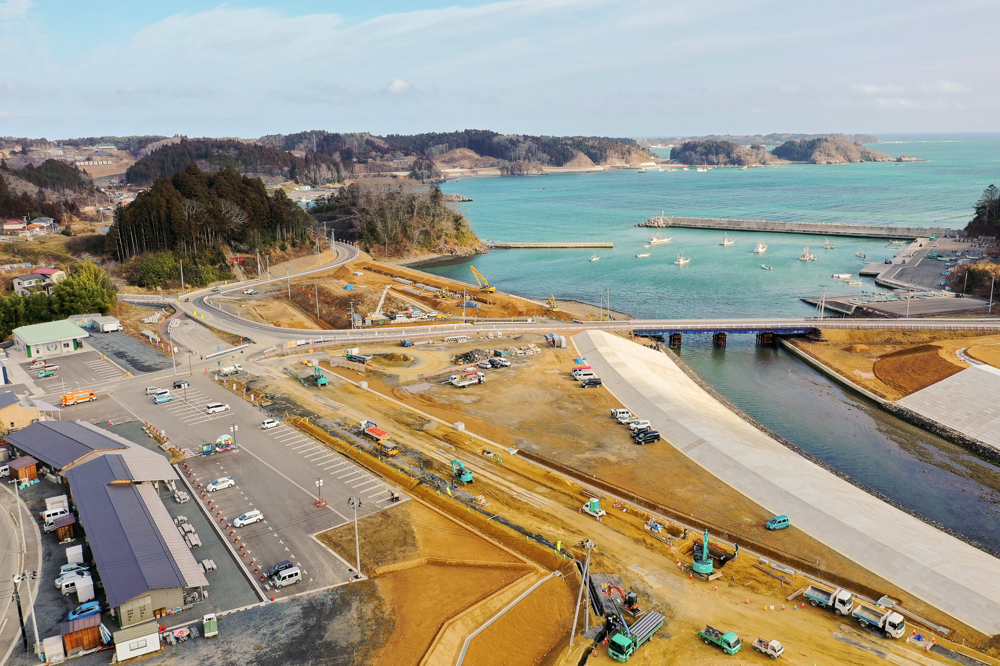海沿いの工事現場を空から撮影した写真で、水辺の近くにはトラックやショベルカーなどの建設機材が点在し、橋が水域をまたいで陸地をつなぎ、背景には桟橋に停泊する小型船と森林に覆われた丘、そして広がる海が見える写真
