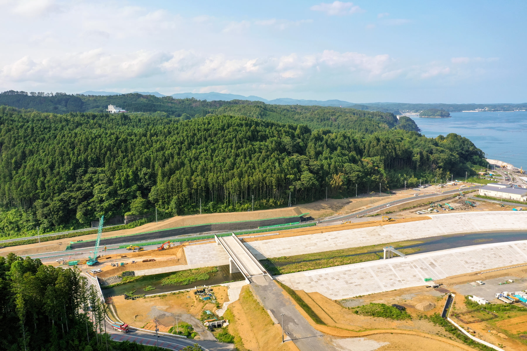 手前に橋と堤防のような構造物がある大規模な工事現場が広がり、背景には緑の森林に覆われた丘があり、右側には海岸線と海が見え、空には雲が少し浮かんでいる沿岸地域の開発風景の写真