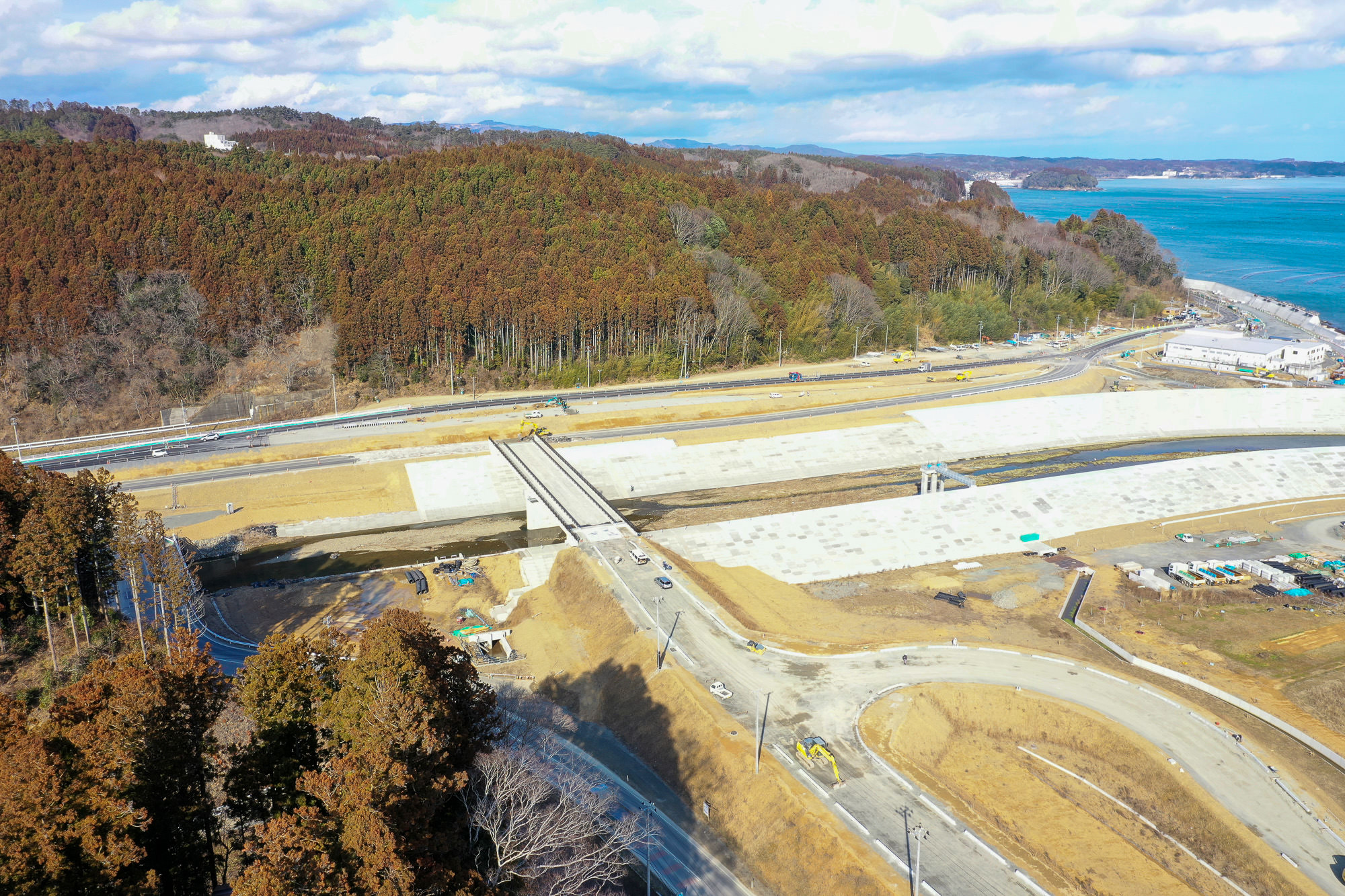 森林と海に近い場所にある工事現場を空から撮影した写真で、道路に架かる橋の上には複数の車両や建設機材が見え、周囲には開発やインフラ整備のために整地された土地が広がる写真