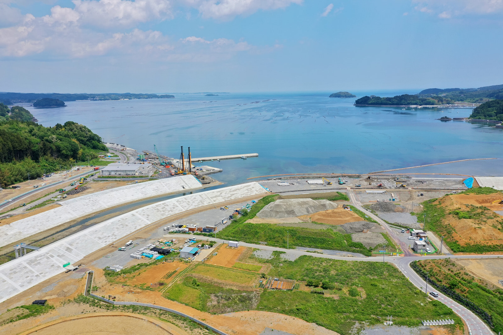 海岸沿いで複数の白い構造物が建設中の工事現場を写した写真で、これらは防潮堤や堤防と思われ、周囲には工事用の機材や資材が点在し、一部は草地で一部は掘削された土地が広がり、背景には穏やかな海と遠くに小さな島々や陸地が見え、空には雲が浮かぶ海岸工事の写真