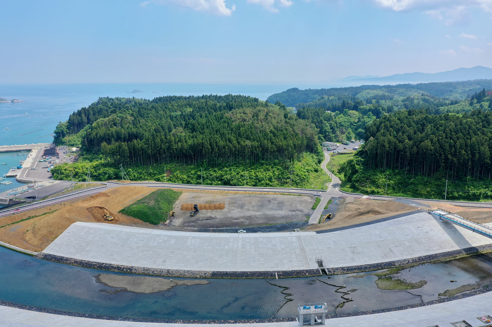 手前に大きなコンクリート構造物が広がり、これは防潮堤またはダムと思われ、背後には密集した緑の森林が広がり、左側には船が停泊する小さな港があり、背景には海と遠くの丘や山々が見える海沿いの地域の写真