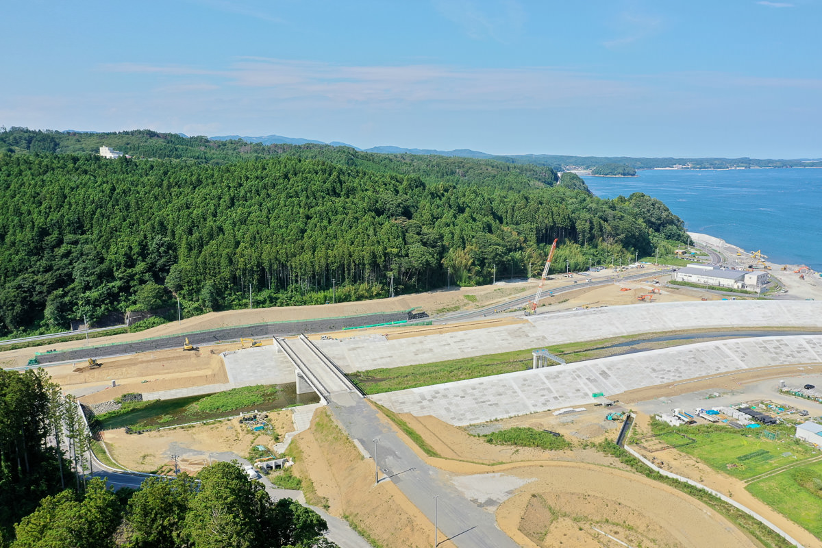 森林と海に近い場所にある工事現場で、中央には建設途中の道路か橋の構造物があり、周囲には建設資材や機材が散在し、背景には濃い緑の木々と右側に海岸線が見え、空は晴れて薄い雲が浮かんでいる工事風景の写真