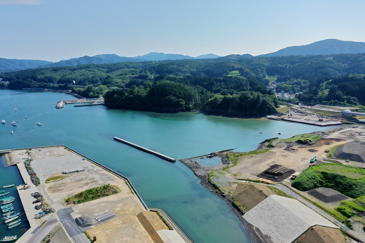 海に囲まれた沿岸地域の空撮で、左側には桟橋と船が並び、右側には開発中または整備された区域が広がり、背景には森林に覆われた丘や山が見え、空は青く澄んでいる海辺の風景の写真
