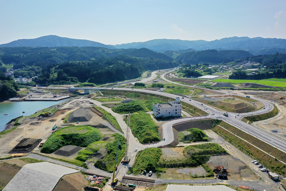 手前に工事現場が広がり、道路や通路、緑地が点在し、中央付近には白い建物があり、背景には緑のある丘や山、点在する建物が見え、空は晴れて雲がほとんどない開発中の地域の写真