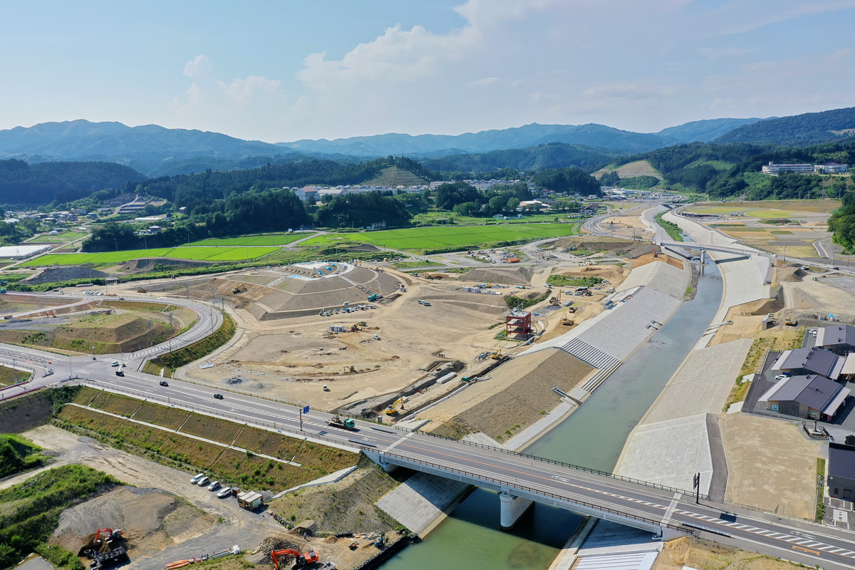 川または運河に架かる橋を中心とした風景で、周囲には造成作業が行われており、土木工事や機械が見られ、道路や通路が整備されつつあり、背景には山々と緑が広がっている開発中の地域の写真