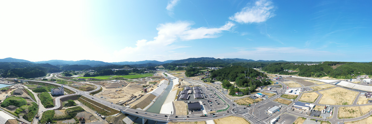 都市的な要素と農村的な要素が混在する風景の広角写真で、手前には道路や建物、工事中の区域があり、中央には川が流れ、その両岸には構造物や緑の畑が広がり、背景には丘や山が見え、空は青く晴れて雲が少し浮かんでいる写真